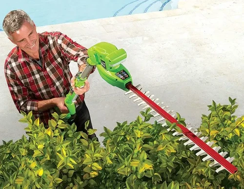  אדם משתמש במגזמת גובה טלסקופית נטענת מבית Greenworks לגיזום שיחים גבוהים ליד בריכה, עם להב ארוך וראש מתכוונן לחיתוך בזוויות שונות.