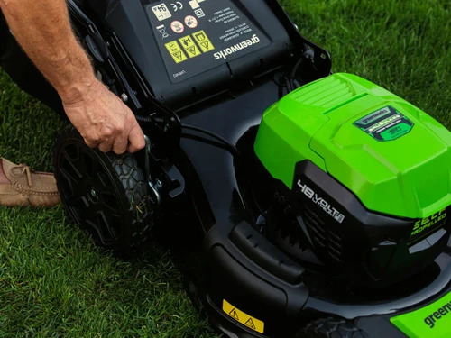 אדם מכוון את גובה הגלגל במכסחת דשא נטענת Greenworks 48V עם מנוע Brushless והנעה עצמית, באמצעות ידית כיוון מרכזית. המכסחת מוצבת על דשא ירוק ועבה.