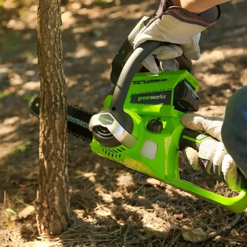 כריתת גזע עץ דק באמצעות מסור שרשרת נטען Greenworks 24V; אדם עם כפפות עבודה מפעיל את המסור בשטח חוץ מיוער.
