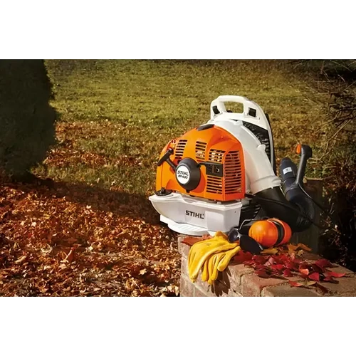 מפוח גב בנזין STIHL BR 430 מונח על קיר לבנים לצד ציוד מגן בעונת הסתיו בגינה מלאה בעלים