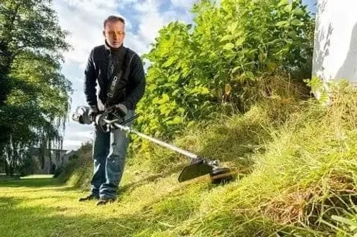 אדם משתמש בחרמש משולב EGO לגיזום עשב גבוה בשטח גינה - עבודה קלה ונוחה עם רצועת נשיאה.