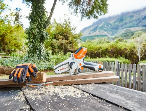 מסור שרשרת קומפקטי STIHL GTA 26 מונח על משטח עבודה בגינה, לצד כפפות עבודה ועץ לביקוע, עם רקע ירוק של צמחייה והרים.