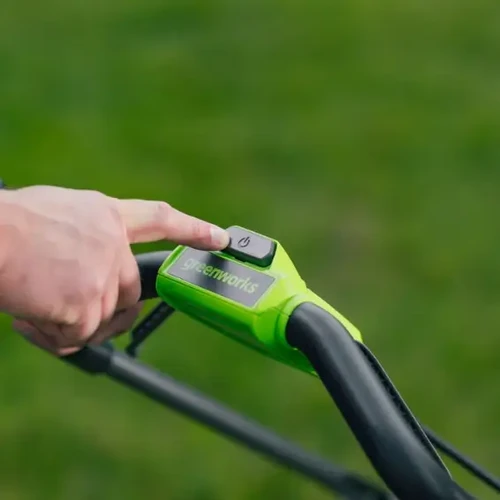 תקריב על ידית אחיזה ירוקה של מכסחת דשא של Greenworks, המראה אצבע לוחצת על כפתור ההפעלה/כיבוי