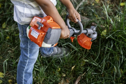 ילד משחק עם חרמש צעצוע Husqvarna Toy Trimmer בגינה, כולל אפקטים קוליים, תאורת LED וידית אחיזה נוחה לחוויית משחק מציאותית ומהנה