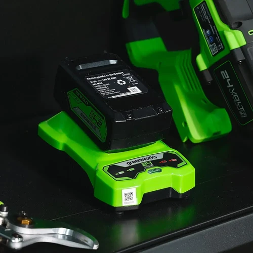 מטען Greenworks 24V במהלך טעינת סוללת ליתיום, לצד כלי עבודה נטען מסדרת 24V בסביבת עבודה מקצועית.