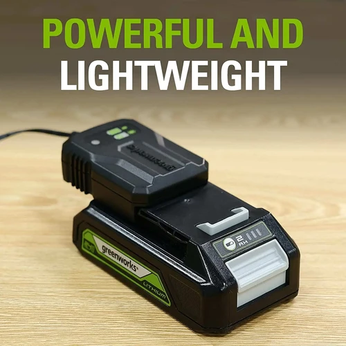 סוללת Greenworks 24V 2Ah עם מטען קומפקטי 2A במהלך טעינה על משטח עבודה, עיצוב קל משקל וקומפקטי עם חיווי מצב טעינה מובנה