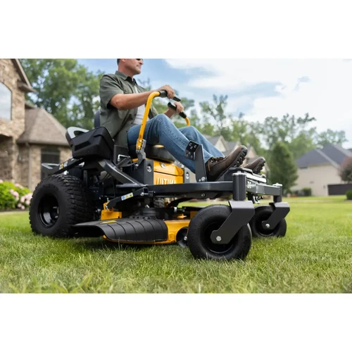 מבט צדי נמוך על טרקטורון כיסוח Cub Cadet Z2-42 בפעולה על דשא, עם מפעיל יושב, ברקע בית מגורים