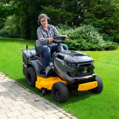 אישה מפעילה טרקטורון כיסוח דשא Cub Cadet LT2 R92 בגינה, לובשת אוזניות מגן ומשקפיים, מבט צדי