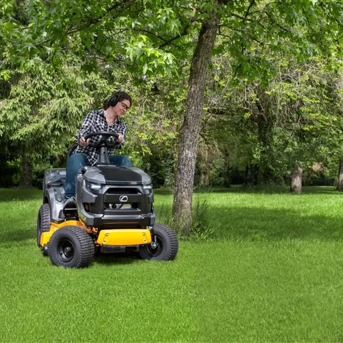 טרקטורון כיסוח דשא Cub Cadet LT2 R92 בשימוש על מדשאה גדולה ליד עץ, מפעילה יושבת על הכלי, מבט חזיתי-צדי